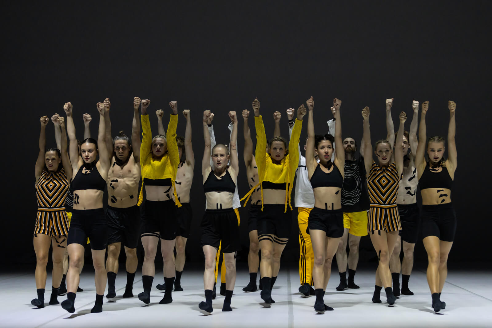 A group of 16 dancers, in two rows of 8 march towards the audience, their hands clenched into fists, their arms punching straight up into the air.