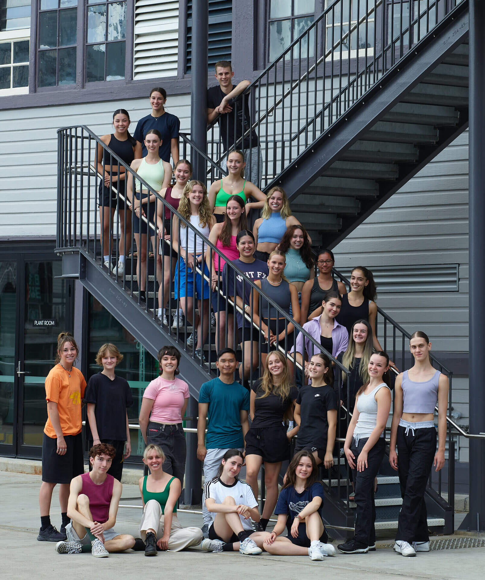 The Pre-Professional Year consort of 60 dancers gathers on the spiral staircase outside the Harbourside home of Sydney Dance Company. They are all smiling into the camera.