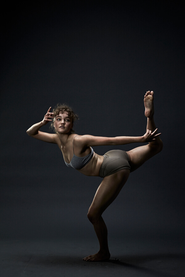 A female dancer balances on one leg with her back left arching up behind her.