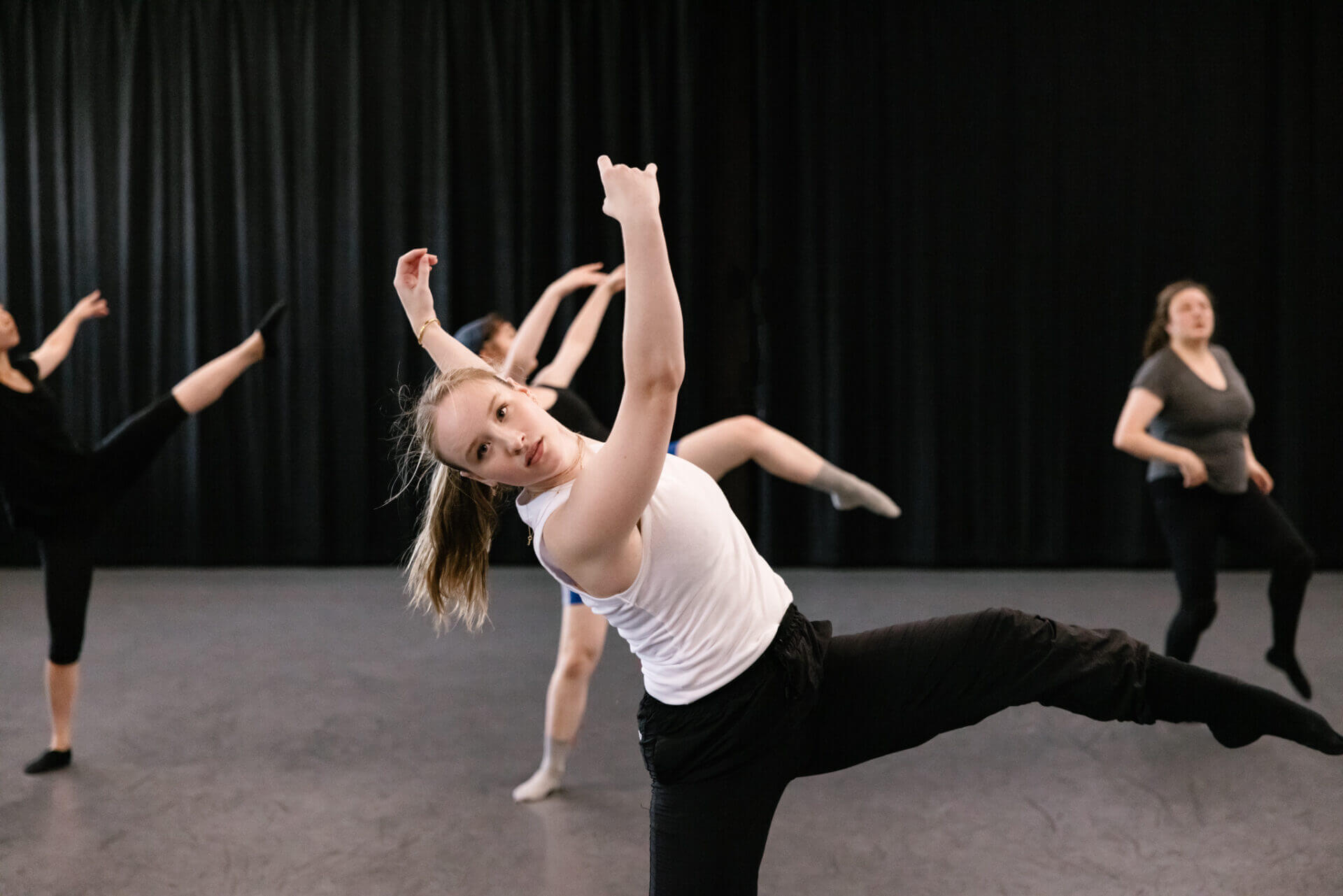 Young dancer in the studio leaning to their right with arms extended upwards and left leg lifted at a 90 degree angle with a bent knee and pointed toe.