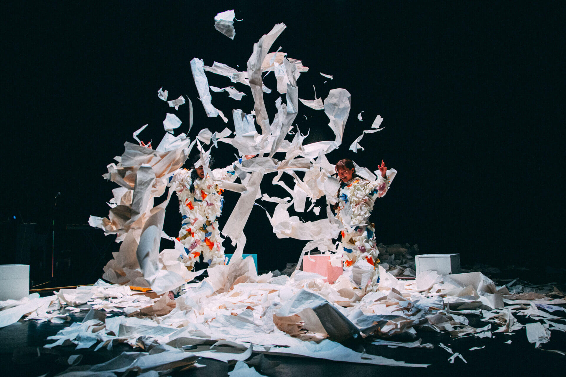 Two performers dressed in paper outfits throwing mounds of paper into the air. Floor is full of paper.