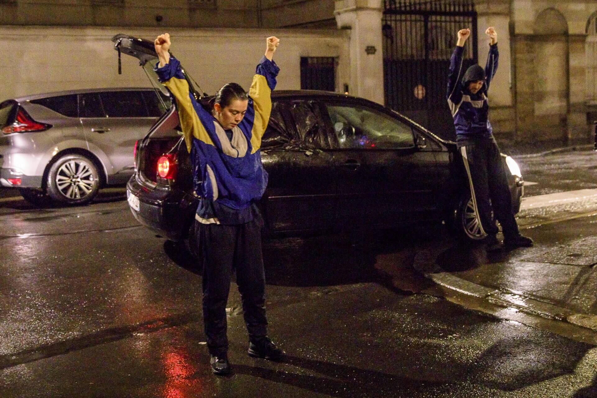 Two people outside a car with their arms outstretched above their heads.